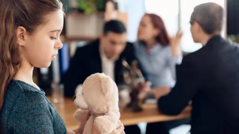 Fillette tenant un ours en peluche, tandis que trois adultes discutent à une table en arrière-plan